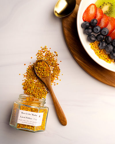 Jar of bee pollen spilled on table with a plate of blueberries, strawberries, and kiwi for pairing.