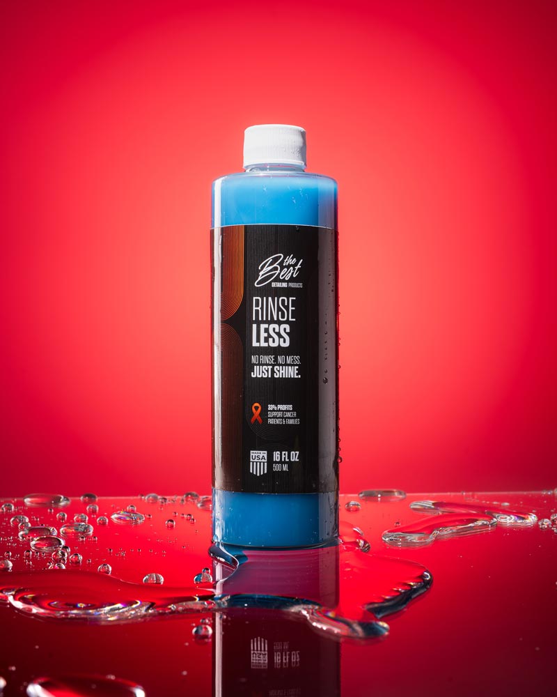 Bottle of rinseless car wash sits on a highly reflective surface with some water showcasing the reflection of the product in front of a bright red background.