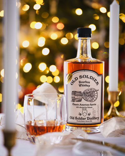 Bottle of old soldier whiskey sits on a stylized table with Christmas lights on a tree in the background