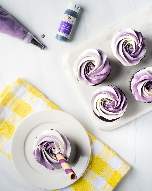 Seattle product photography - Ube Overload. Lifestyle image from the top of Ube Overload cupcakes on a bright table surface with a piping tool full of ube frosting and a lone ube cupcake on a bright yellow napkin.
