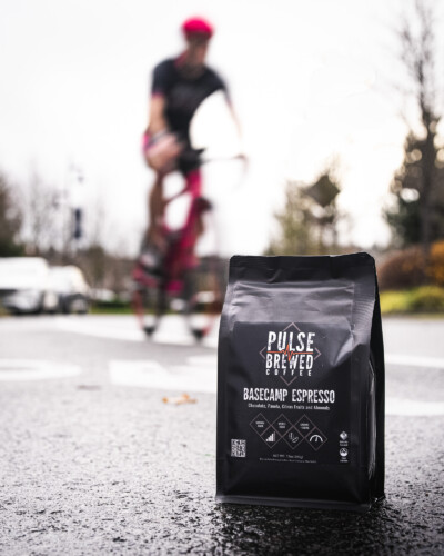 Using a slower shutter to blur the bike behind the mug and coffee beans emphasizes movement, connecting Pulse Brewed coffee to action, adventure, and an active, engaged community. Pulse Brewed Basecamp Espresso Coffee on the road with bicyclist passing by