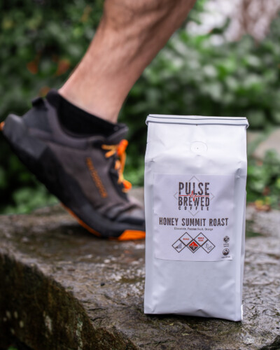 By capturing movement on the trail behind the branded coffee, this image links Pulse Brewed to active living, making the brand relatable and purpose-driven. Pulse Brewed Honey Summit Roast Coffee on the trail with trail runner passing by