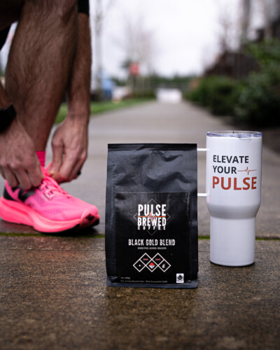By combining a branded mug, coffee beans, and a runner preparing their shoes, this image connects Pulse Brewed coffee to active lifestyles and real moments, reinforcing the brand’s story and purpose. Pulse Brewed Black Gold Blend Coffee on the sidewalk with runner lacing shoes for run