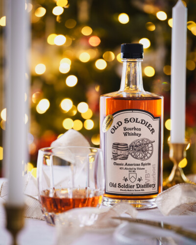 By placing candles in the foreground and background, we created layers that lead the viewer through this holiday scene, bringing focus to Old Soldier Vanilla Bean Whiskey while keeping the setting warm and festive. Old Soldier Vanilla Bean bourbon whiskey sits on a table with a glass of poured bourbon whiskey showcasing a bright, elegant christmas spirit and highlighting the perfect pairing of the product with the holidays