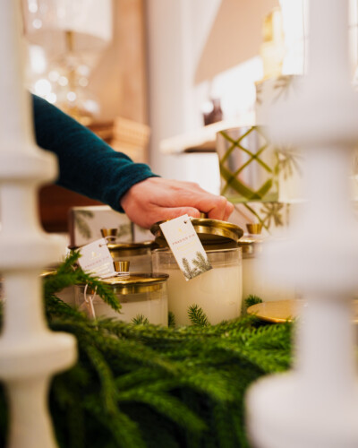 By framing the hand lifting the lid off the candle between two heavily blurred foreground stick candles, we create depth, guide the viewer’s eye, and add a sense of intimacy and warmth to the scene. A candle is opened amongst the House & Hazel holiday decor