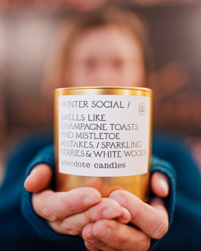 Hands holding the candle draw the viewer in, creating warmth and connection while keeping the person softly in the background. This subtle human element adds emotion and context to the scene. A candle is held out for a winter social