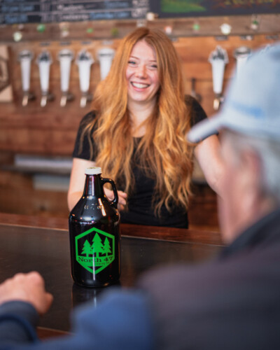 By focusing on the bartender passing the growler, this image illustrates the brand’s personality and commitment to customer experience, using human interaction to make the brand more relatable and memorable. A growler is passed across the bar at North 47 Brewing Co.