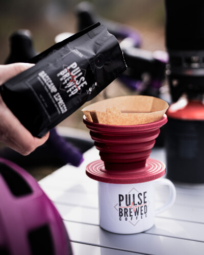 By including a human element, the photo invites the viewer into the moment — a cup of Pulse Brewed coffee on a camping table with a Jetboil — highlighting the brand’s active lifestyle identity. Bile leaning against table with Jetboil and helmet, while Pulse Brewed coffee is poured into a drip filter to make a on-the-go coffee