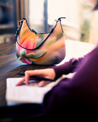 Showcase your product in use to share the quality and style for your customers. A purse from Sally's Blooming Bags sits on a table as a person writes notes in a journal