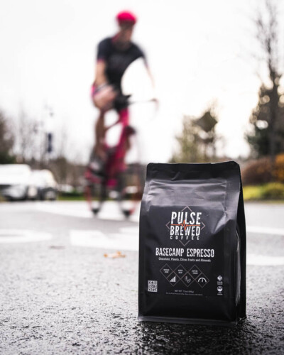 Seattle product photography for Pulse Brewed Coffee in Black Diamond, WA. Basecamp Espresso and coffee beans sit on the road while a cyclist blurs past in the background - slow-shutter lifestyle content linking this Greater Seattle Area coffee brand to cycling and active outdoor adventure. Seattle Product Photography - Pulse Brewed Basecamp Espresso on Road with Motion-Blurred Cyclist Passing - Active Lifestyle Coffee - Black Diamond, WA - Elemental Brand Photography