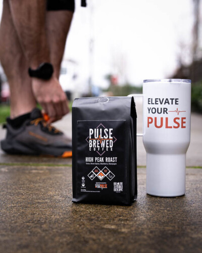 Seattle product photography for Pulse Brewed Coffee in Black Diamond, WA. High Peak Roast coffee bag and branded mug on the sidewalk alongside a runner lacing their shoes - relatable active lifestyle product photography for a community-driven Greater Seattle Area coffee brand. Seattle Product Photography - Pulse Brewed High Peak Roast with Running Shoes and Coffee Beans on Sidewalk - Pacific Northwest Active Coffee Brand - Black Diamond - Elemental Brand Photography