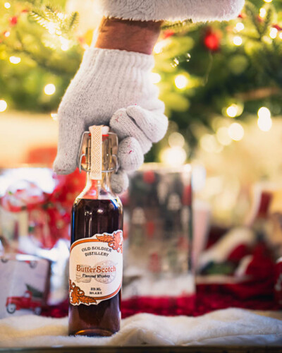 Seattle Product Photography for Old Soldier Distillery in Tacoma, WA. Best0selling Butterscotch Whiskey delivered to the Christmas tree in a bright lit scene for seasonal marketing and social media imagery Seattle Product Photography - Old Soldier Distillery - Tacoma, WA - Seasonal Santa Clause Butterscotch Whiskey - Elemental Brand Photography