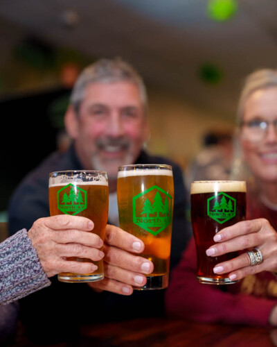 Seattle Restaurant photography for North 47 Brewing Co. in Tacoma. Three branded beer glasses held out in a lively toast with customers enjoying the experience - brand content capturing the community spirit of a Pacific Northwest craft brewery. Seattle Restaurant Photography - North 47 Brewing Co. Beer Glasses Held Out- Tacoma Craft Brewery - Elemental Brand Photography