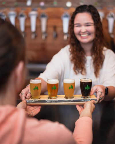 Seattle Restaurant Photography for North 47 Brewing Co. in Tacoma, WA. An over-the-shoulder moment captures a bartender handing off a beer flight - approachable, lifestyle-forward brand content for a Greater Seattle Area craft brewery. Seattle Restaurant Photography - Over-the-Shoulder Shot of Beer Flight Being Passed at North 47 Brewing Co. - Tacoma, WA - Elemental Brand Photography