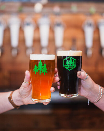 Seattle Restaurant Photography for North 47 Brewing Co. in Tacoma. Dark and amber beers in branded glassware held in the foreground, taproom bar visible behind - evergreen social media content connecting product to place for a community-driven craft brewery. Seattle Restaurant Photography - North 47 Dark and Amber Beers Held Up with Taproom Bar Behind - Tacoma Brewery - Elemental Brand Photography