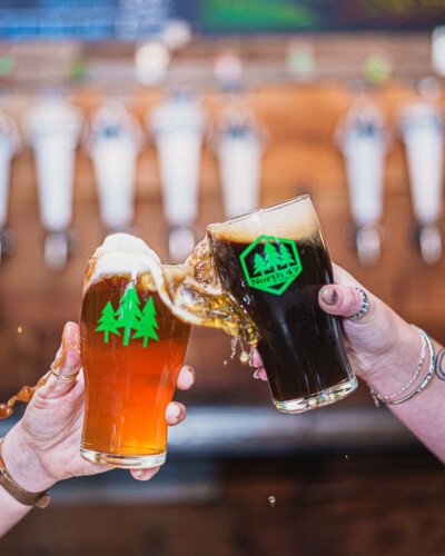Seattle Restaurant photography for North 47 Brewing Co. in Tacoma. Two branded beer glasses collide in a lively toast, spilling over the sides - movement-forward brand content capturing the community spirit of a Pacific Northwest craft brewery. Seattle Restaurant Photography - North 47 Brewing Co. Beer Glasses Clinking in Celebration - Tacoma Craft Brewery - Elemental Brand Photography