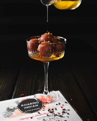 Seattle Restaurant Photography for Roaring Indian Plates & Pours in Maple Valley, WA. Gulab jamoon in a warm spotlight of light with the fresh, sweet syrup being poured on the desert. Social media and marketing content created to showcase the quality and experience of the local, authentic Indian restaurant. Seattle Restaurant Photography - Roaring Indian Plates & Pours - Syrup pour on Gulab Jamoon - Maple Valley, WA - Elemental Brand Photography
