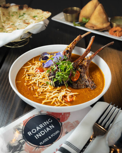 Seattle Restaurant Photography for Roaring Indian Plates & Pours in Maple Valley, WA. Gourmet confit with fresh naan and samosas cooked in the kitchen of the James Beard finalist with warm lighting spotlighting the main dish. Social media content created to showcase the beautifully plated curry plate at the local pacific northwest restaurant. Seattle Restaurant Photography - Roaring Indian Plates & Pours - Curried Lamb Confit - Maple Valley, WA - Elemental Brand Photography